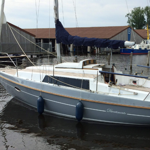 Auf dem Bild liegt eine graue Neptun 26 im Wasser. Das Segelboot hat eine Scheuerleiste aus Teakholz, aufgesetzte uv-beständige Fenster und eine Schiebelukgarage mit elektronischen Windrichtungsanzeiger und Nasa Clipper Duett. Die Plicht und das Deck sind mit Teakholzbelag ausgestattet.